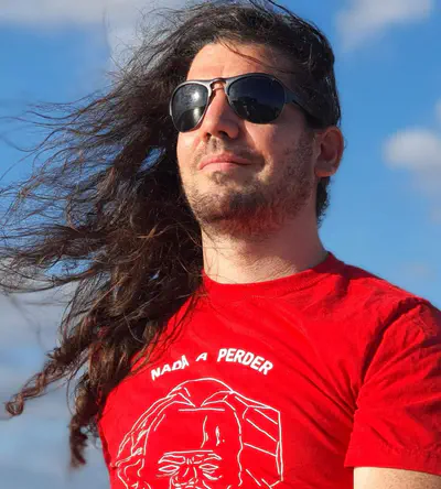 Foto de rosto do Giovanni Bassi. Ele é branco e de cabelos compridos, olha para a esquerda, está com os cabelos soltos, óculos escuros e uma camiseta vermelha, onde se lê 'nada a perder'.